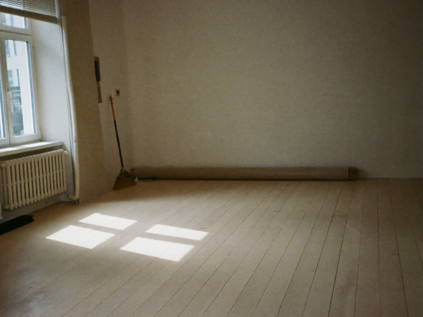 Ein leerer Raum mit hellen Dielen; am Fenster ein Heizkörper, in der Ecke ein Besen und ein zusammengerollter Teppich an der Wand, auf dem Boden ein Lichtfleck